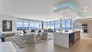 A modern living room with a large window overlooking the sea, a kitchen with stainless steel appliances, and a cozy seating area with a rug and a sofa. - Ocean Front with Rooftop Pool Access (Destin)