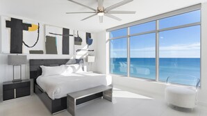 A modern bedroom with a large bed, a ceiling fan, a nightstand, a bedside table, and a view of the ocean. - Ocean Front with Rooftop Pool Access (Destin)