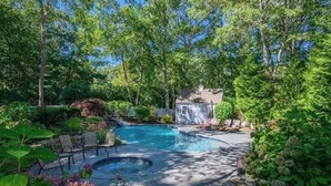A backyard with a swimming pool, stone seating, and lush greenery. - Private Deep Water Dock, heated pool and hot tub RPMS005 (Marstons Mills)