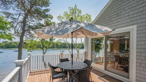 A wooden deck with a table and chairs, an umbrella, and a view of a lake with boats. - Private Deep Water Dock, heated pool and hot tub RPMS005 (Marstons Mills)