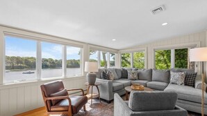 A spacious living room with a large gray sectional sofa, a brown leather armchair, and a small side table. Large windows provide a view of a body of water and greenery. - Private Deep Water Dock, heated pool and hot tub RPMS005 (Marstons Mills)