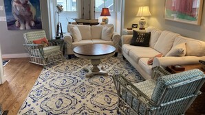 Un salon avec un tapis à motifs, un canapé blanc, un fauteuil à motifs, une table basse ronde et un tableau représentant un lapin.