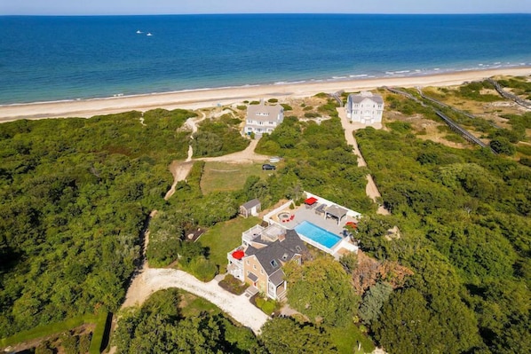 An aerial view of a coastal property with multiple buildings, a swimming pool, and a beach in the background. - Secluded Escape w/ Heated Pool and Private Beach Access RPAM001 (East Sandwich)