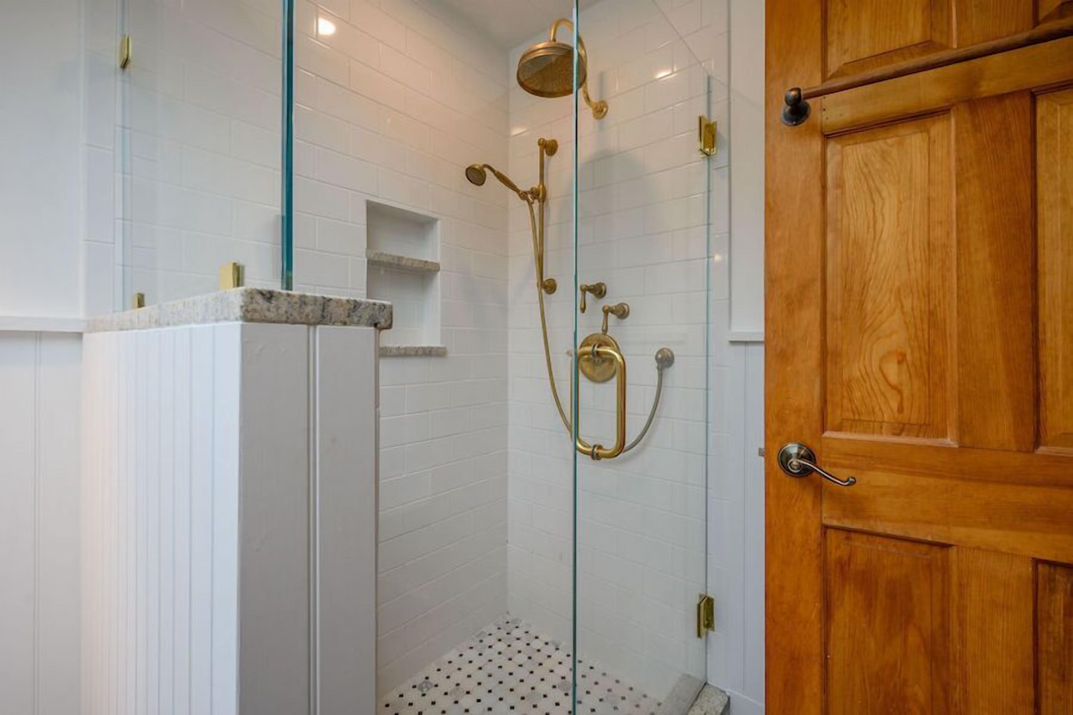 A glass-enclosed shower with a gold showerhead and controls, a wooden door to the right, and white tiled walls.