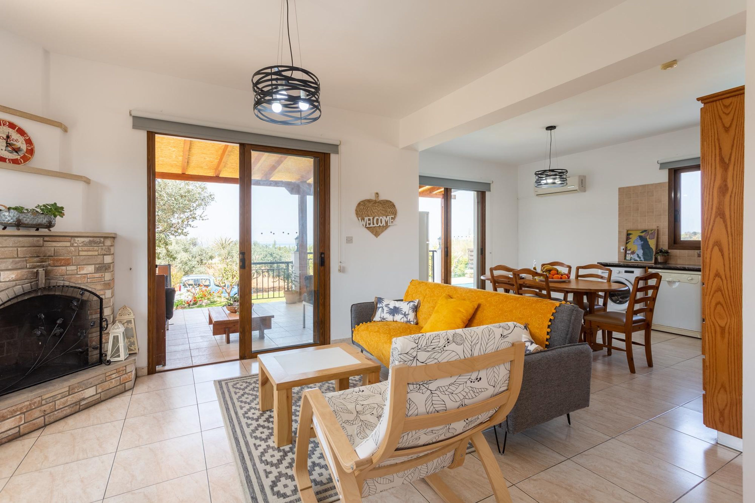 A spacious living area with a fireplace, a sofa with yellow cushions, a dining table, and a kitchen in the background.
