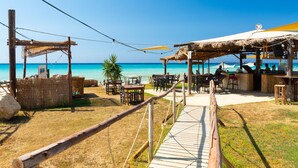 A beachside restaurant with outdoor seating, a thatched-roof bar area, and a wooden walkway leading to the beach.