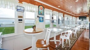 A restaurant with wooden paneling, white chairs, round tables, and large windows offering a view of the sea.
