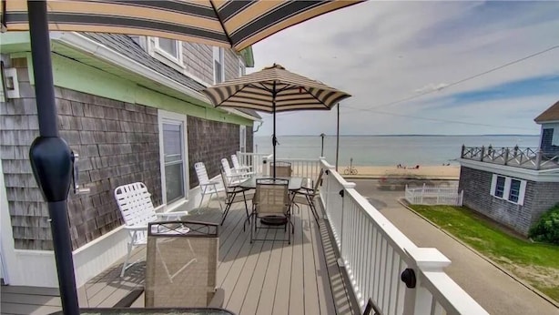 A wooden deck with outdoor furniture, a glass table, and a beach view.