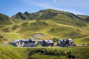 A hotel with multiple buildings, set against a backdrop of green hills and mountains. - 100m from resort center, 2nd floor, jacuzzi, hammam, balcony, parking, tv, ski locker, 59m² (Germ)