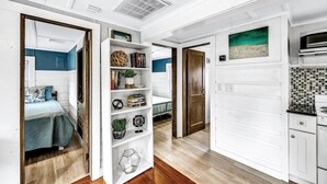 A modern interior with a hallway leading to a bedroom with a blue accent wall, a white shelving unit with various items, and a kitchen area with a tiled backsplash.