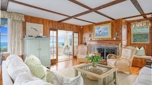 A spacious living room with wooden walls and a fireplace, furnished with white sofas and wicker chairs, and large windows offering a view of the outdoors. - Craigville Long Beach Beachfront rental home. On private beach. RPMS029 (Centerville)
