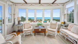 A room with a view of the sea, featuring white wicker furniture, a wooden coffee table, and a window with curtains. - Craigville Long Beach Beachfront rental home. On private beach. RPMS029 (Centerville)