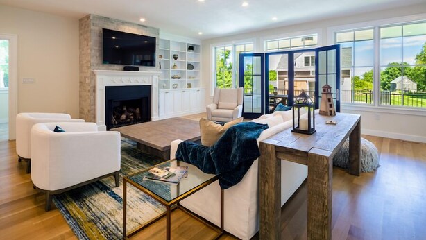A modern living room with a fireplace, white sofas, a coffee table, and a view of the outdoors through large windows. - Dazzling High End Home with Heated Pool, Spa and Guest House RPAM013 (East Dennis)