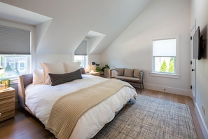 A bedroom with a large bed, a desk, a chair, a TV, and a window with blinds.
