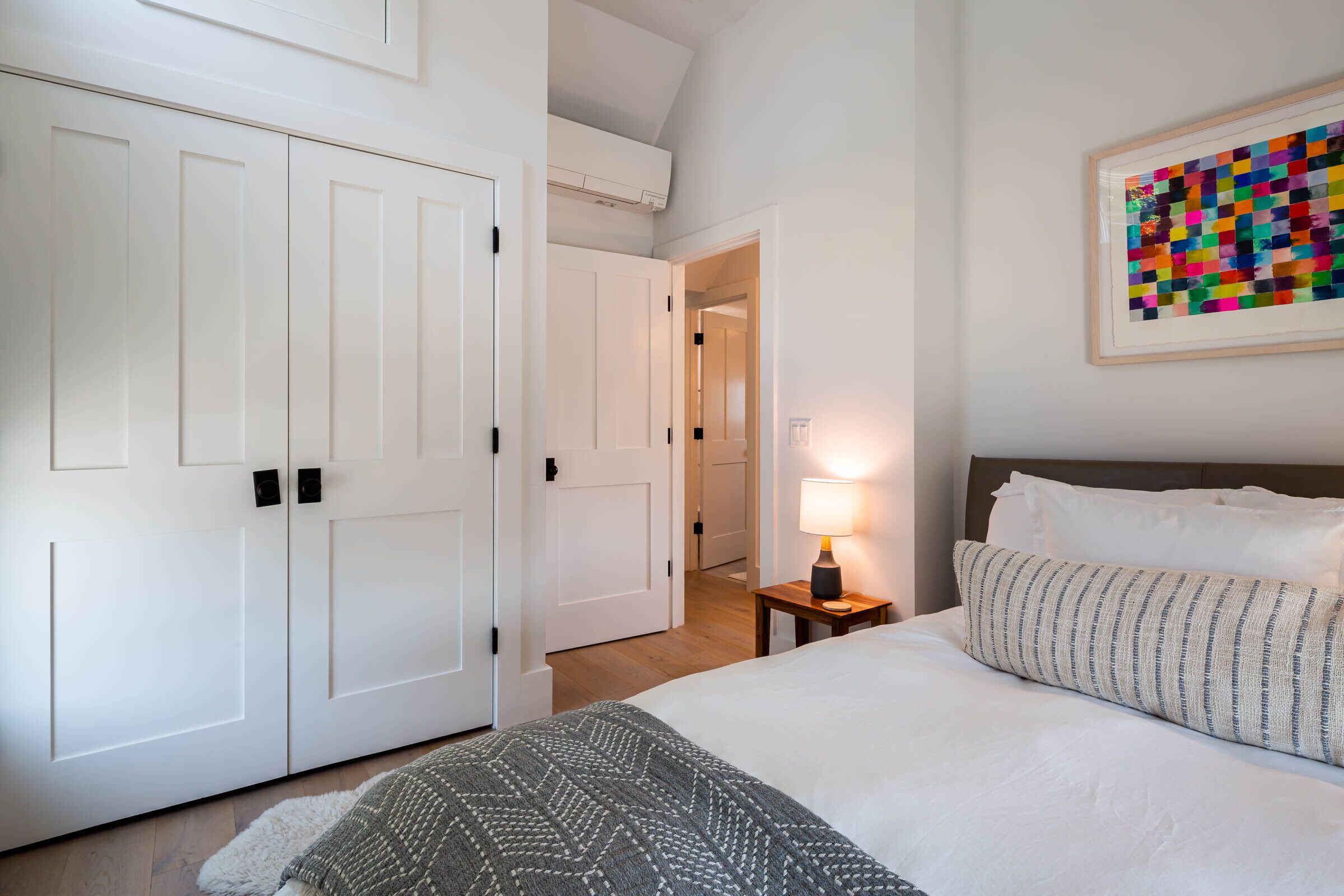 A bedroom with a bed, a nightstand with a lamp, a framed artwork, and a white wardrobe.