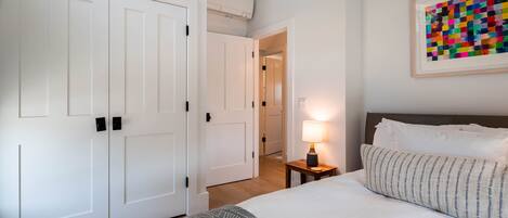 A bedroom with a bed, a nightstand with a lamp, a framed artwork, and a white wardrobe.