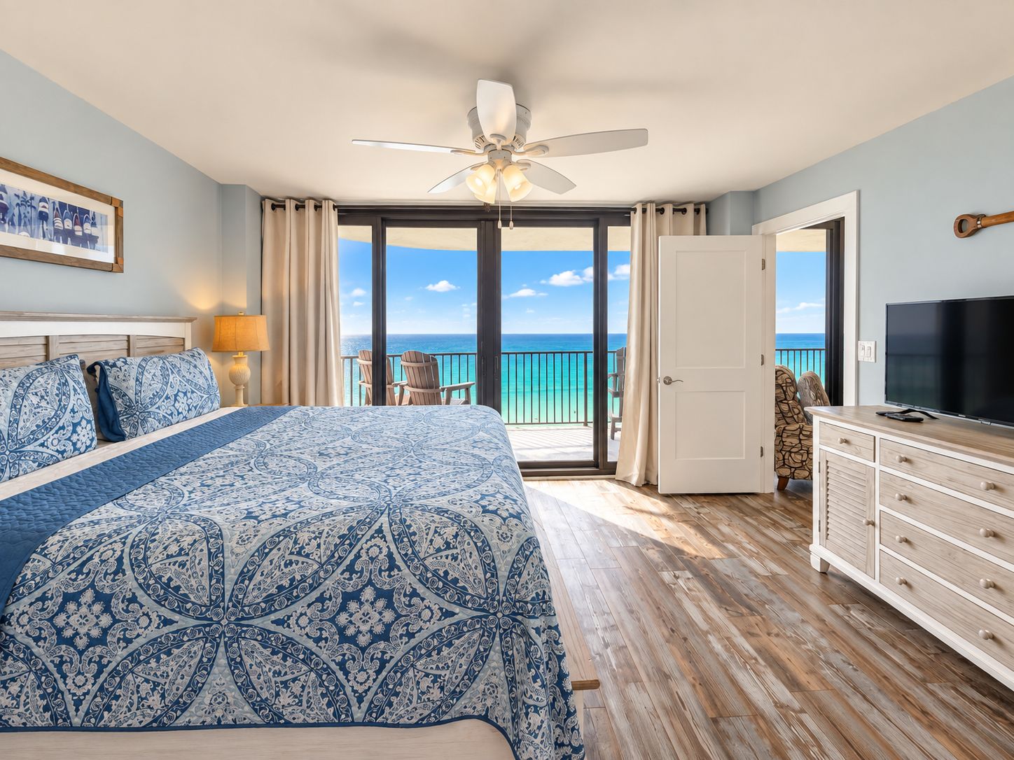 Quarto de hotel com cama, TV, varanda com vista para o oceano e ventilador de teto.