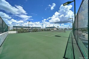 Una cancha de tenis con superficie verde, rodeada por una cerca de malla metálica y bajo un cielo azul despejado con nubes.