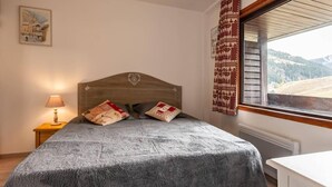 A bedroom with a wooden headboard, a grey bedspread, two pillows, a bedside lamp, and a window with a view of a mountainous landscape. - 20m from ski slopes, 2nd floor, balcony, fireplace or stove, tv, 48m², Méribel-Mottaret (Méribel - Les Allues)