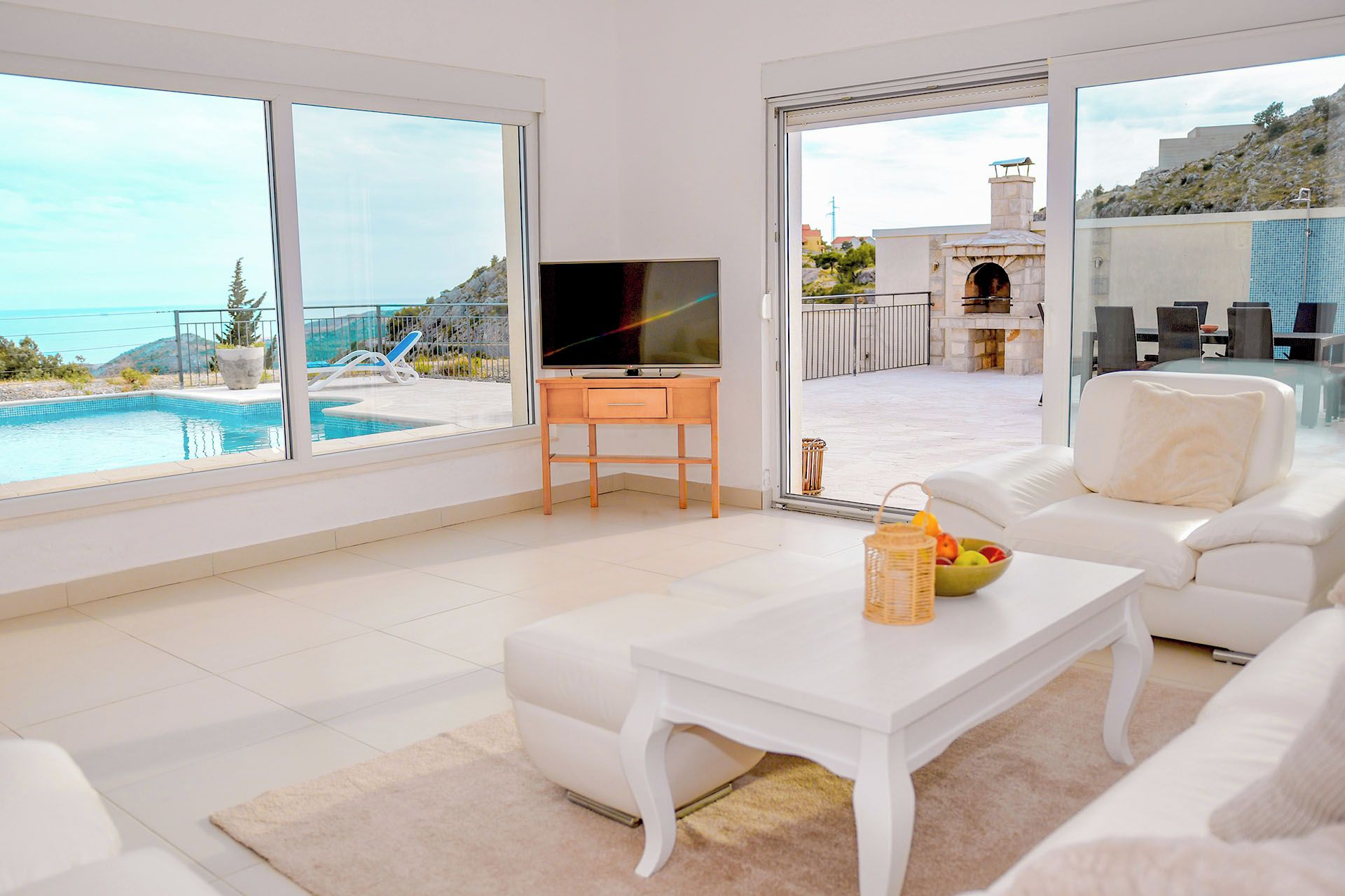 A modern living room with a pool view, a flat-screen TV, and a dining area with a view of the pool and outdoor seating.