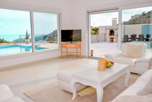 A modern living room with a pool view, a flat-screen TV, and a dining area with a view of the pool and outdoor seating.