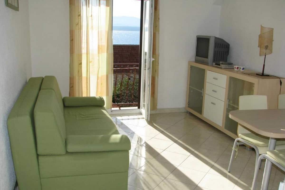 A room with a green sofa, a small dining table with chairs, a TV on a cabinet, and a view of the sea through a balcony.