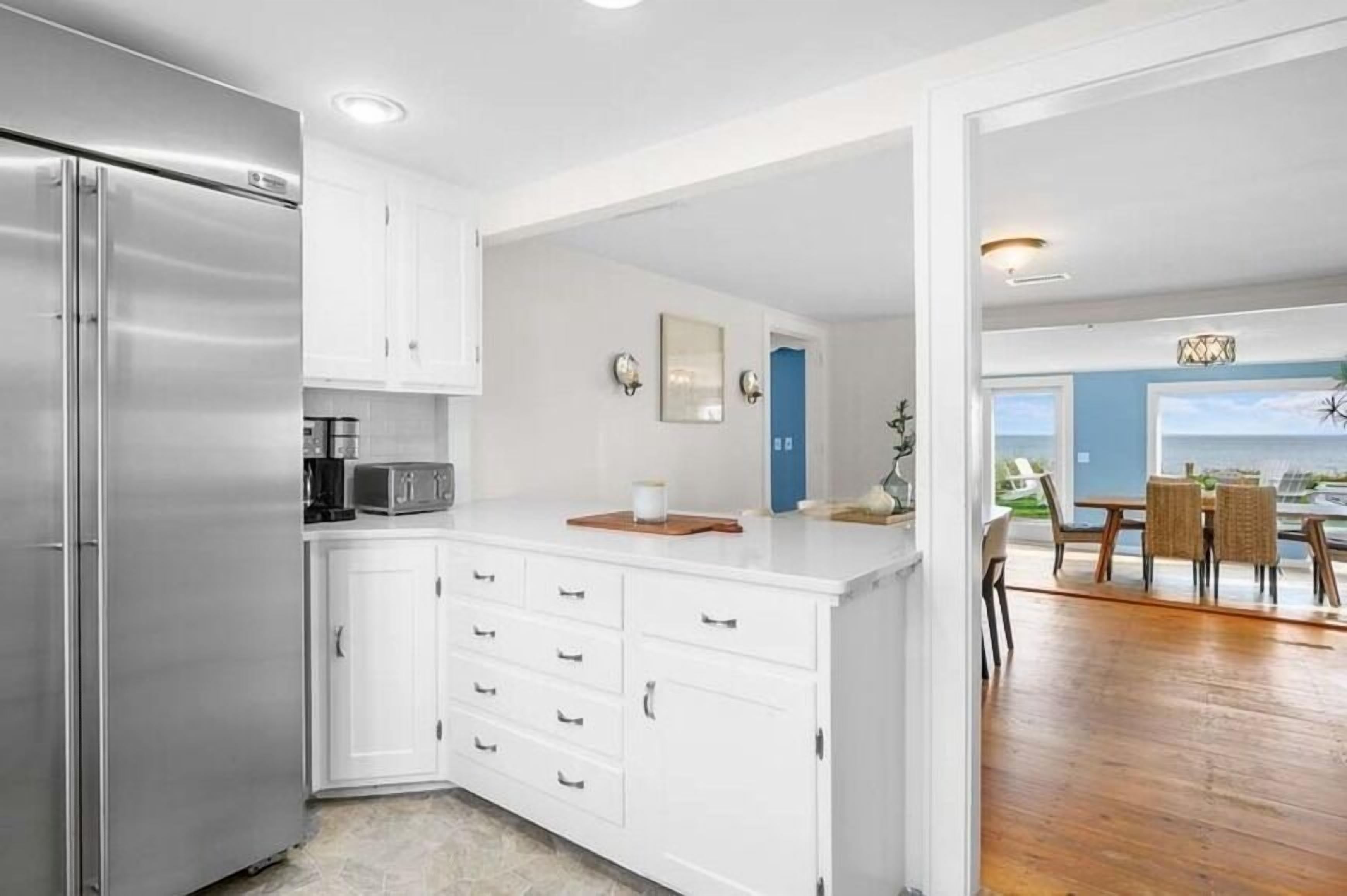 A modern kitchen with white cabinetry, a stainless steel refrigerator, and a microwave. There is a dining area with a table and chairs visible through an open doorway.
