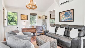 A spacious living room with a large black leather sofa, grey armchairs, and a wicker ottoman. A wooden side table with a lamp and decorative items is placed near a window. A tall grandfather clock and a framed map are on the wall. - Time and Tide Cottage - A Barnstable Village Retreat RPAM020 (Barnstable)