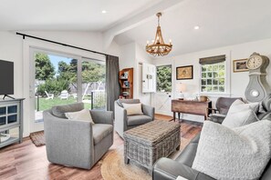 A spacious living room with a large window, a chandelier, and a wooden coffee table. - Time and Tide Cottage - A Barnstable Village Retreat RPAM020 (Barnstable)