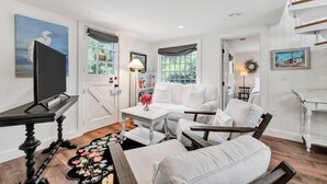 A spacious living room with a white sofa set, a flat-screen TV, and a dining area with a table and chairs. - Time and Tide Cottage - A Barnstable Village Retreat RPAM020 (Barnstable)