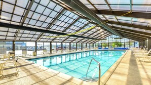 An indoor swimming pool with clear glass ceilings, sunlit pool area, and lounge chairs along the sides.