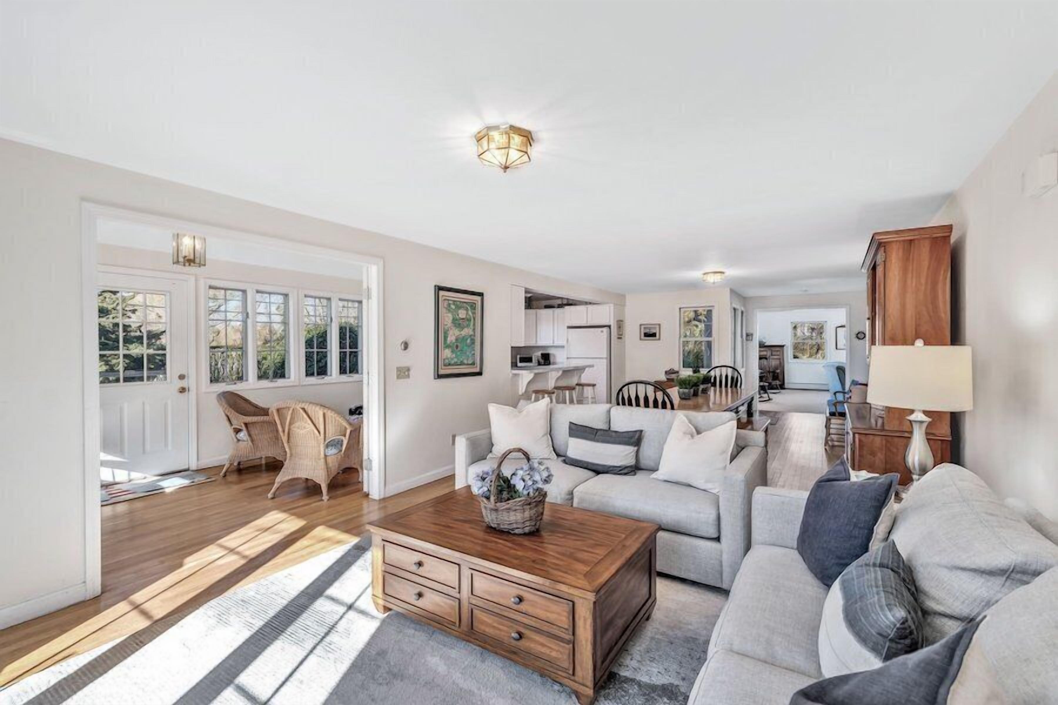 Ein geräumiges Wohnzimmer mit Couchtisch aus Holz, grauen Sofas, Korbstühlen und einer Küche im Hintergrund.