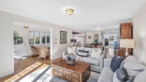 A spacious living room with a wooden coffee table, grey sofas, wicker chairs, and a kitchen in the background. - Private Beach Front Chatham Rental Close To Everything RPAM054 (Chatham)