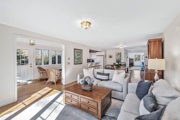 A spacious living room with a wooden coffee table, grey sofas, wicker chairs, and a kitchen in the background.