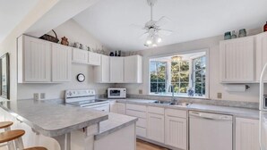 A modern kitchen with white cabinetry, a central island with stools, a built-in oven, microwave, and dishwasher, and a large window providing natural light. - Private Beach Front Chatham Rental Close To Everything RPAM054 (Chatham)