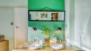 Une table à manger dressée avec des assiettes, des couverts et des verres, entourée de chaises, avec une plante en pot et un tableau encadré au-dessus.