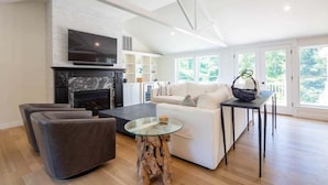 A modern living room with a fireplace, a flat-screen TV, a white sofa, and a glass coffee table. - Elevate Your Cape Stay: Chatham Luxury with Pool & Beach RPAM026 (Chatham)
