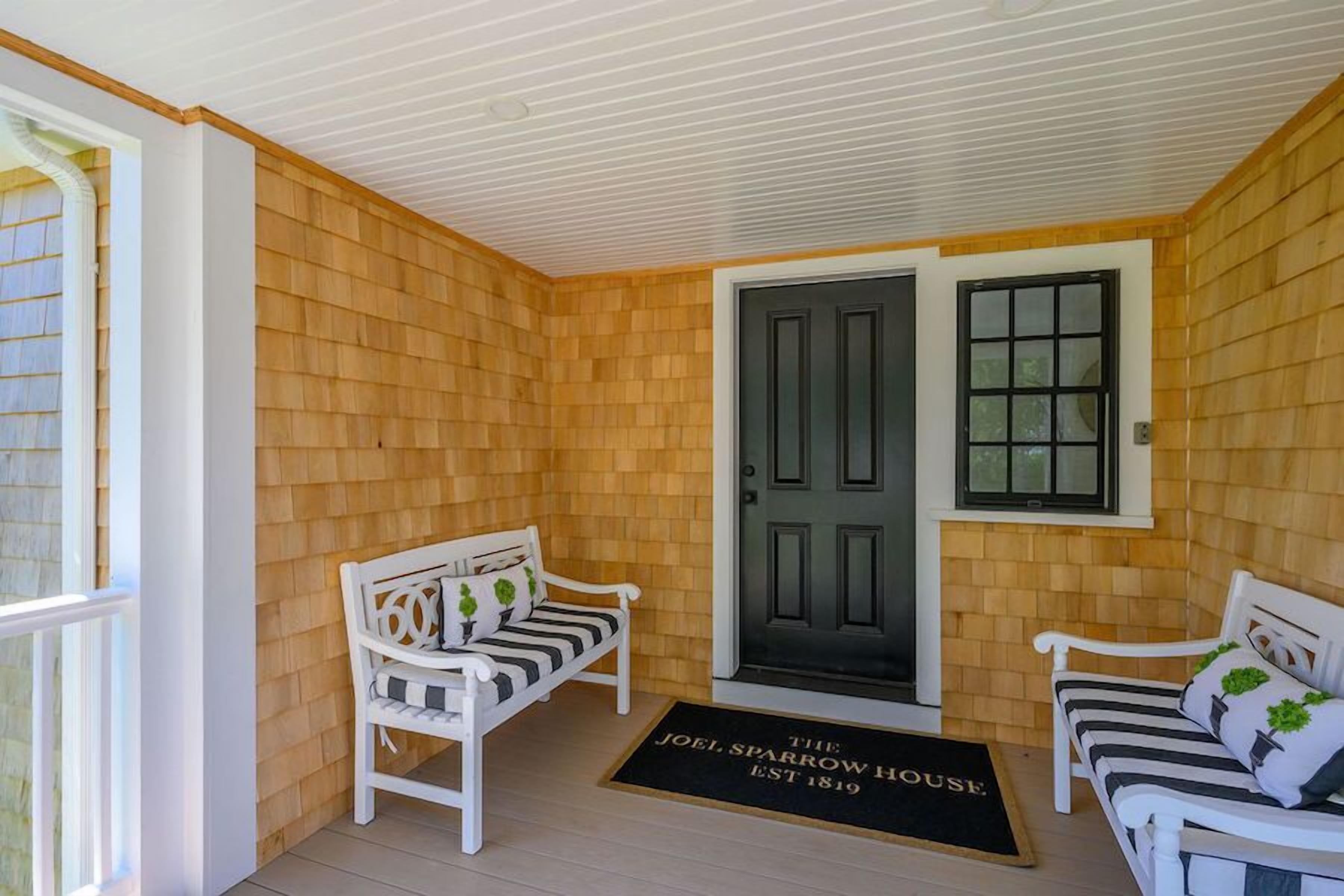 A cozy porch with two white benches, a black door, and a doormat reading "The Joel Sparrow House EST 1819."