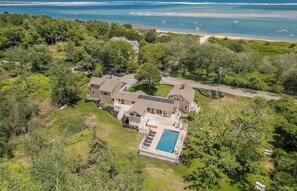An aerial view of a large house with a swimming pool, surrounded by dense greenery and a beach. - Elevate Your Cape Stay: Chatham Luxury with Pool & Beach RPAM026 (Chatham)
