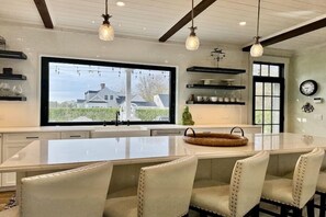 Una cocina moderna con una isla grande, armarios blancos, iluminación colgante y vista a una casa a través de un ventanal.