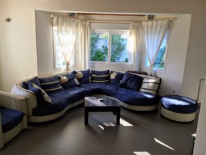 Un salon moderne avec un canapé d’angle aux lignes courbes, une petite table basse et un pouf rond. Il y a deux fenêtres avec des rideaux, et le sol est carrelé.