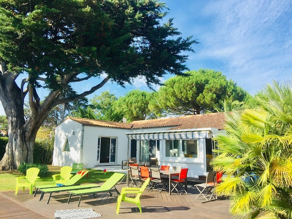 A white building with a tiled roof, outdoor seating area with green and orange chairs, and a large tree in the foreground.