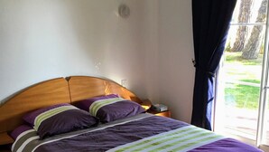 Un dormitorio con cabecera de madera, cama con ropa de cama morada y rayas verdes, mesita de noche y ventana con cortinas.