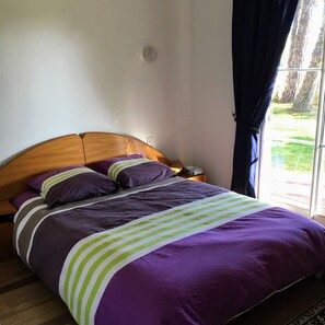 Ein Schlafzimmer mit einem hölzernen Kopfteil, einem Bett mit violetter Bettwäsche und grünen Streifen, einem Nachttisch und einem Fenster mit Vorhängen.