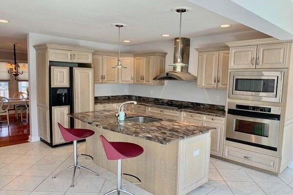 A kitchen with light wood cabinets, a granite countertop, stainless steel appliances, and two red bar stools. - Spacious Falmouth Retreat with Private Pool RPAM072 (Falmouth)