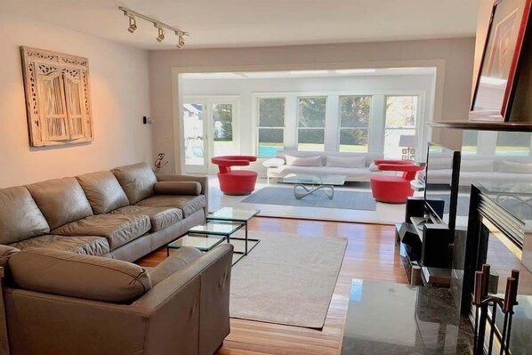 A modern living room with a leather sofa, glass coffee table, and red accent chairs. There is a fireplace and a large window offering a view of the outdoors. - Spacious Falmouth Retreat with Private Pool RPAM072 (Falmouth)
