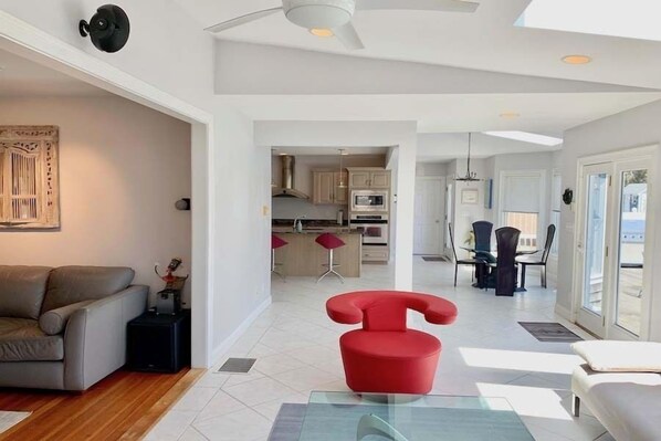 Een moderne woonkamer met een glazen salontafel, een rode fauteuil en een grijze bank. Er is een keuken zichtbaar op de achtergrond met een eethoek.