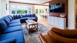 Una sala de estar moderna con un sofá modular, una mesa de centro y un sillón de cuero. Un televisor de pantalla plana está montado en la pared, y se aprecia un comedor y un espacio exterior.