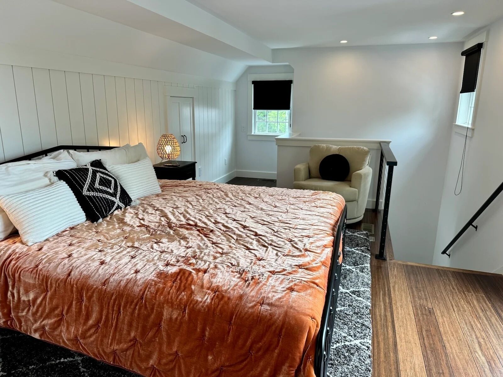 A bedroom with a large bed, a nightstand, a chair, a window, and a staircase.