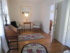A room with a wooden floor, a floral rug, a sofa, two chairs, a table with a lamp, and a painting on the wall.